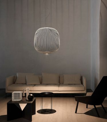 A minimalist living room with the Spokes pendant lamp by Foscarini as an elegant focal point. The lamp's delicate metal struts cast soft patterns of light on the walls and ceiling, giving the room a light, sculptural effect. A light-colored sofa, dark accents, and natural materials create a calm, modern atmosphere. Die filigranen, metallischen Streben der Leuchte werfen sanfte Lichtspiele an Wand und Decke und verleihen dem Raum eine leichte, skulpturale Wirkung. Helles Sofa, dunkle Akzente und natürliche Materialien schaffen eine ruhige, moderne Atmosphäre.