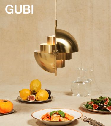 An elegant Multi-Lite pendant lamp by GUBI in brushed brass hangs above a set table. Beneath it are plates of pasta, fresh figs, and lemons, as well as a glass carafe—a stylish scene that combines design and enjoyment.