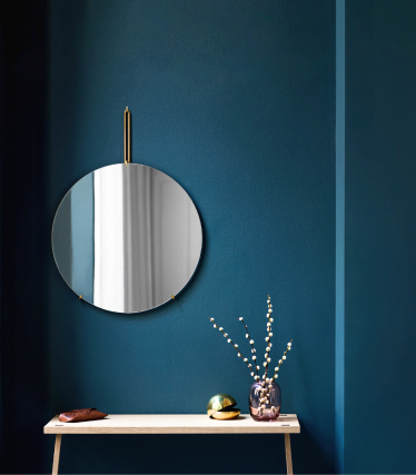 A round wall mirror with a brass hanging detail mounted on a deep blue wall. Below it stands a light wooden bench decorated with a small vase of branches, a decorative object, and a leather accessory. The scene feels minimalist, elegant, and Scandinavian in style.