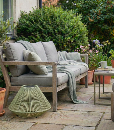 Cosy wooden bench with cushions and blanket on a garden terrace, surrounded by plants and a small side table.