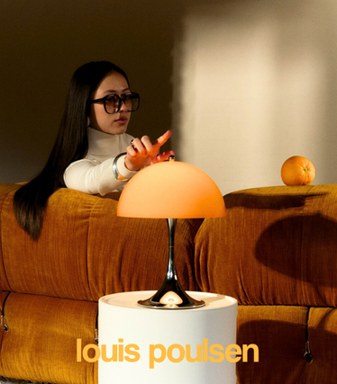 Woman with sunglasses touches an orange Louis Poulsen table lamp from the new Panthella collection. The light stands on a white side table in front of an orange-coloured sofa, in warm, soft light.