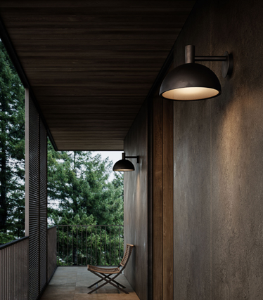 Narrow, covered outdoor corridor with dark wood and concrete walls. Two black Nordlux wall lights provide warm light along the corridor, with a single chair at the end in front of green nature.