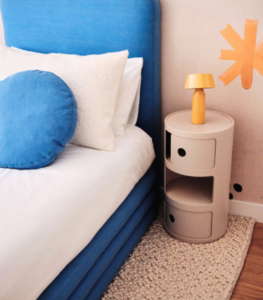A blue upholstered bed with white bedding and a round blue pillow sits beside a small beige cylindrical bedside table topped with a yellow lamp, against a wall with an orange abstract sunburst design and a textured rug on a wooden floor.