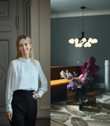 Portrait of designer Sofie Refer next to a luminous pendant light from Nuura in a stylish interior with floral accents.